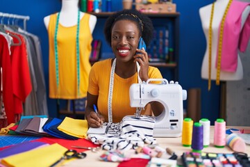 African american woman tailor talking on smartphone writing on notebook at sewing studio