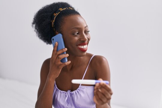 African american woman talking on smartphone holding pregnancy test at bedroom