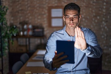 Handsome hispanic man working at the office at night doing stop sing with palm of the hand. warning expression with negative and serious gesture on the face.