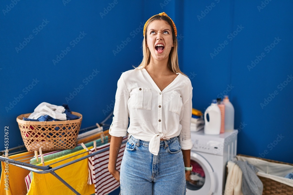 Sticker young blonde woman at laundry room angry and mad screaming frustrated and furious, shouting with ang