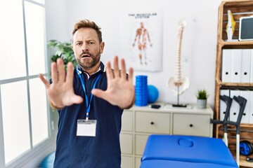 Middle age physiotherapist man working at pain recovery clinic afraid and terrified with fear expression stop gesture with hands, shouting in shock. panic concept.