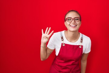 Fototapeta premium Young hispanic woman wearing waitress apron over red background showing and pointing up with fingers number four while smiling confident and happy.
