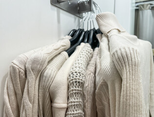 Beige new many stack of soft knitted sweaters hanging on hangers in fitting room in store mall.