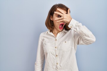 Young beautiful woman standing casual over blue background peeking in shock covering face and eyes...