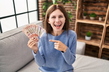 Young brunette woman holding colombian pesos smiling happy pointing with hand and finger - obrazy, fototapety, plakaty
