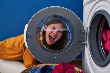 Young brunette woman looking through the washing machine window angry and mad screaming frustrated and furious, shouting with anger. rage and aggressive concept.