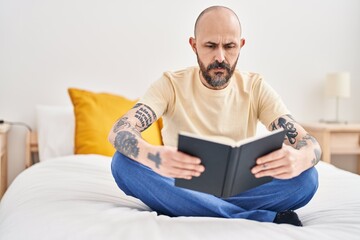 Young bald man reading book sitting on bed at bedroom