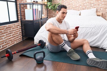 Young hispanic man using smartphone sitting on yoga mat at bedroom