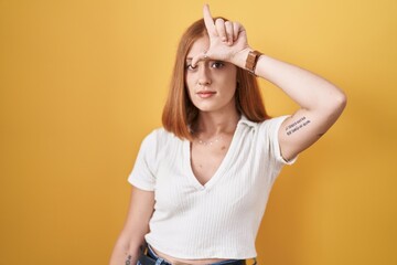 Young redhead woman standing over yellow background making fun of people with fingers on forehead...