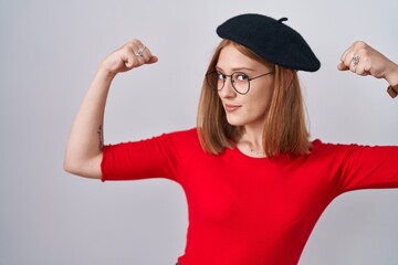 Young redhead woman standing wearing glasses and beret showing arms muscles smiling proud. fitness concept.