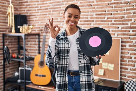 Beautiful african american woman holding vinyl record at music studio doing ok sign with fingers, smiling friendly gesturing excellent symbol