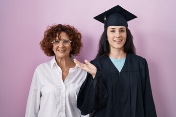 Hispanic mother and daughter wearing graduation cap and ceremony robe smiling cheerful presenting...
