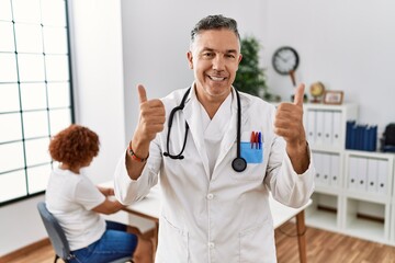 Middle age doctor man at the clinic with a patient success sign doing positive gesture with hand, thumbs up smiling and happy. cheerful expression and winner gesture.