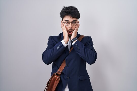 Hispanic man with beard wearing business clothes afraid and shocked, surprise and amazed expression with hands on face
