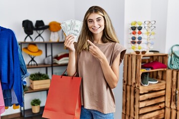 Young blonde woman holding shopping bags and dollar at clothing shop smiling happy pointing with hand and finger