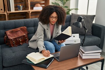 Young african american woman psychologist using laptop and writing book at psychology center