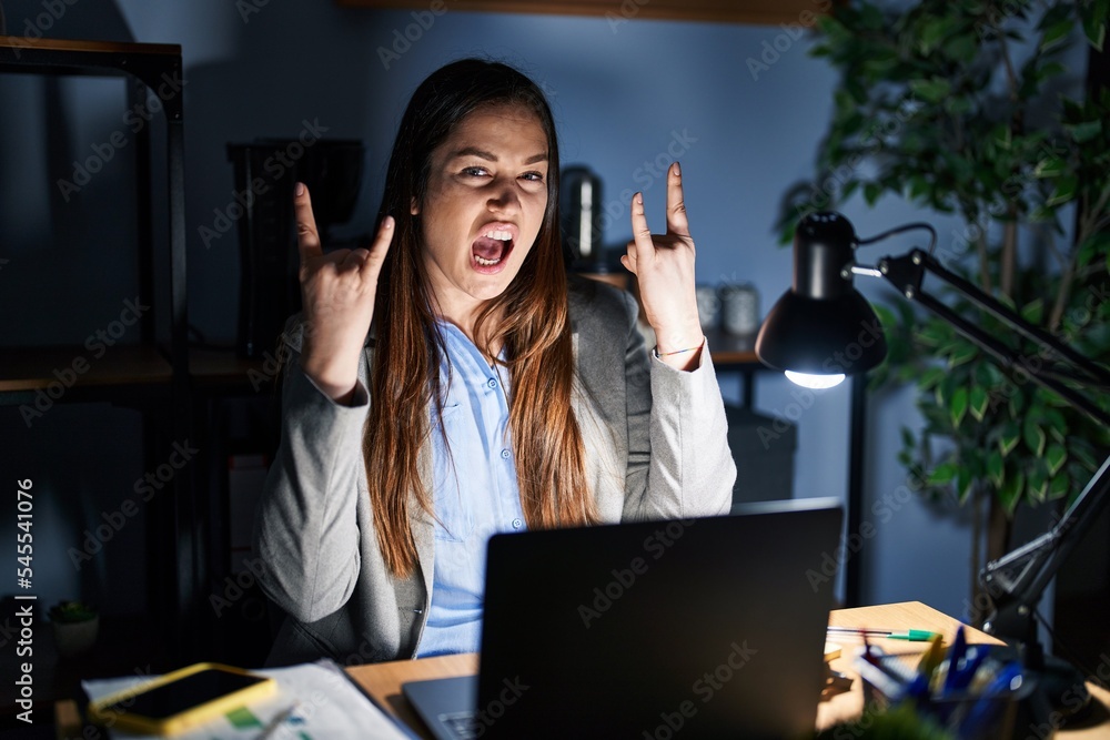 Poster Young brunette woman working at the office at night shouting with crazy expression doing rock symbol with hands up. music star. heavy concept.