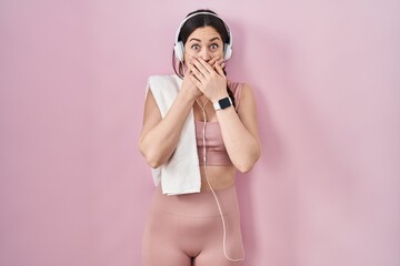 Young brunette woman wearing sportswear and headphones shocked covering mouth with hands for...