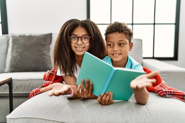 Two siblings lying on the sofa reading a book smiling cheerful offering palm hand giving assistance and acceptance.