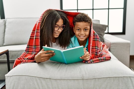 Brother and sister covering with blanket reading book lying on sofa at home