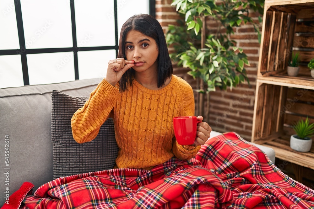 Canvas Prints Young hispanic woman sitting on the sofa drinking a coffee at home mouth and lips shut as zip with fingers. secret and silent, taboo talking