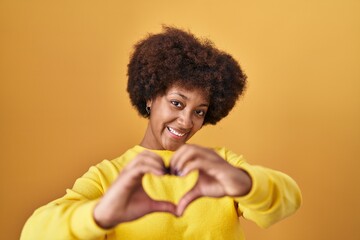 Young african american woman standing over yellow background smiling in love doing heart symbol shape with hands. romantic concept.