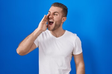 Fototapeta premium Young caucasian man standing over blue background shouting and screaming loud to side with hand on mouth. communication concept.