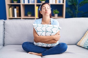 Young hispanic woman hugging pillow sitting on the sofa puffing cheeks with funny face. mouth inflated with air, catching air.