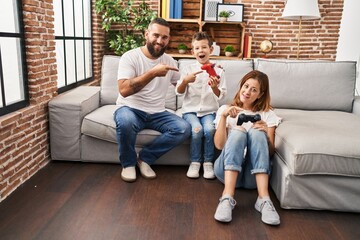 Family of three playing video game sitting on the sofa smiling happy pointing with hand and finger