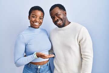 Young african american couple standing over blue background pointing aside with hands open palms...