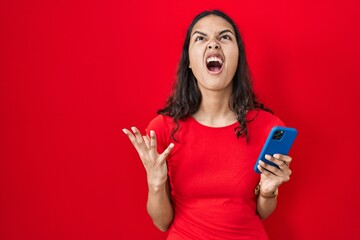 Naklejka premium Young brazilian woman using smartphone over red background crazy and mad shouting and yelling with aggressive expression and arms raised. frustration concept.