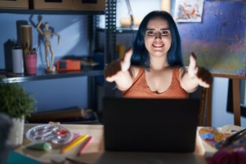 Fototapeta na wymiar Young modern girl with blue hair sitting at art studio with laptop at night approving doing positive gesture with hand, thumbs up smiling and happy for success. winner gesture.