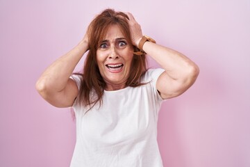 Middle age woman standing over pink background crazy and scared with hands on head, afraid and...