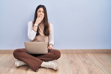 Young brunette woman working using computer laptop sitting on the floor bored yawning tired covering mouth with hand. restless and sleepiness.
