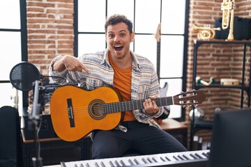 Young hispanic man playing classic guitar at music studio smiling happy pointing with hand and finger - obrazy, fototapety, plakaty