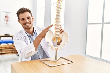 Young hispanic man wearing physiotherapist uniform pointing to anatomical model of vertebral column at clinic