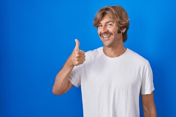 Naklejka premium Middle age man standing over blue background doing happy thumbs up gesture with hand. approving expression looking at the camera showing success.