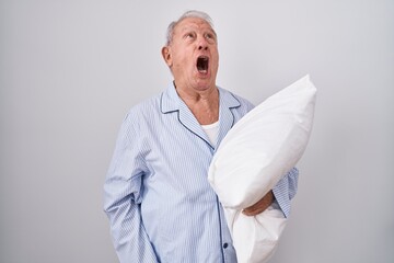 Senior man with grey hair wearing pijama hugging pillow angry and mad screaming frustrated and...