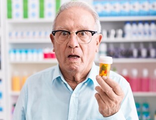 Senior man with grey hair holding pills at the pharmacy scared and amazed with open mouth for...