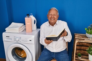 Obraz premium Senior man reading book waiting for washing machine at laundry room