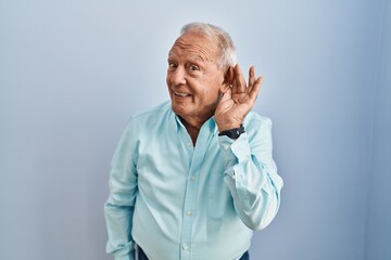 Senior man with grey hair standing over blue background smiling with hand over ear listening an...