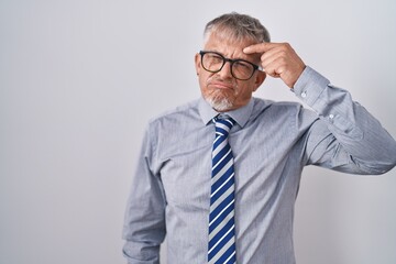 Hispanic business man with grey hair wearing glasses pointing unhappy to pimple on forehead, ugly infection of blackhead. acne and skin problem
