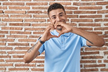 Brazilian young man standing over brick wall smiling in love doing heart symbol shape with hands....