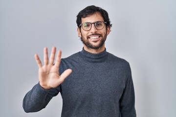 Handsome latin man standing over isolated background showing and pointing up with fingers number five while smiling confident and happy.