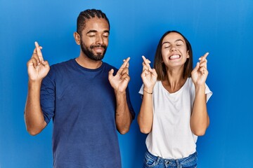 Young hispanic couple standing together gesturing finger crossed smiling with hope and eyes closed....