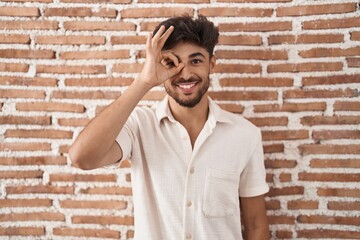 Arab man with beard standing over bricks wall background doing ok gesture with hand smiling, eye looking through fingers with happy face.