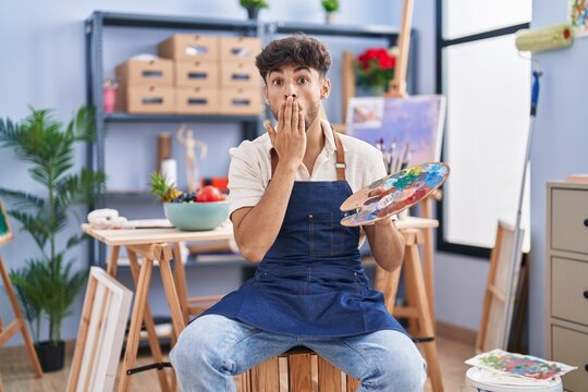 Arab man with beard painter sitting at art studio holding palette covering mouth with hand, shocked and afraid for mistake. surprised expression