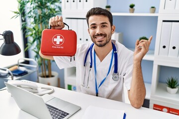 Young hispanic doctor man holding first aid kit smiling happy pointing with hand and finger to the...