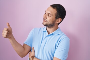 Hispanic man standing over pink background looking proud, smiling doing thumbs up gesture to the side