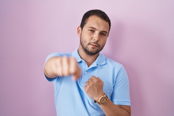 Hispanic man standing over pink background punching fist to fight, aggressive and angry attack, threat and violence